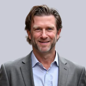 Smiling man in a grey suit with a light blue shirt, set against a neutral background.