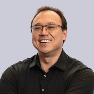 Mike Kenworthy smiles confidently with folded arms, wearing glasses and a black shirt against a light purple background.