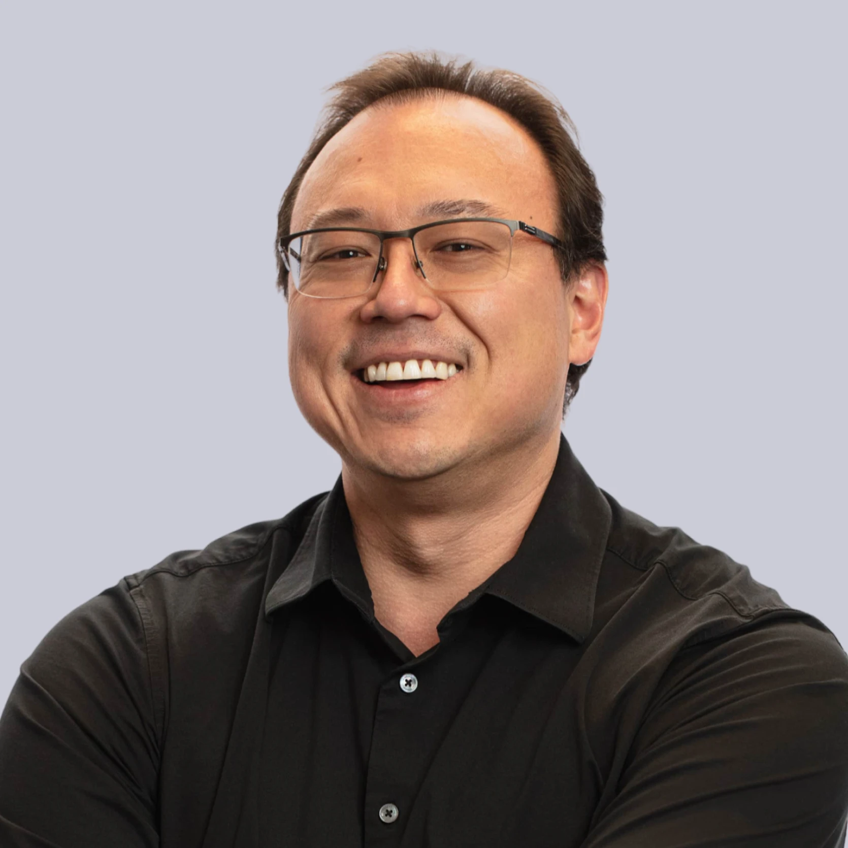 Mike Kenworthy smiles confidently with folded arms, wearing glasses and a black shirt against a light purple background.