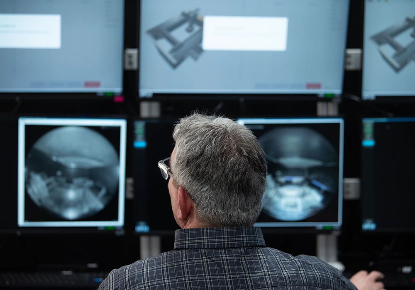 A person monitors multiple computer screens displaying software interfaces and technical images in a control room setup.