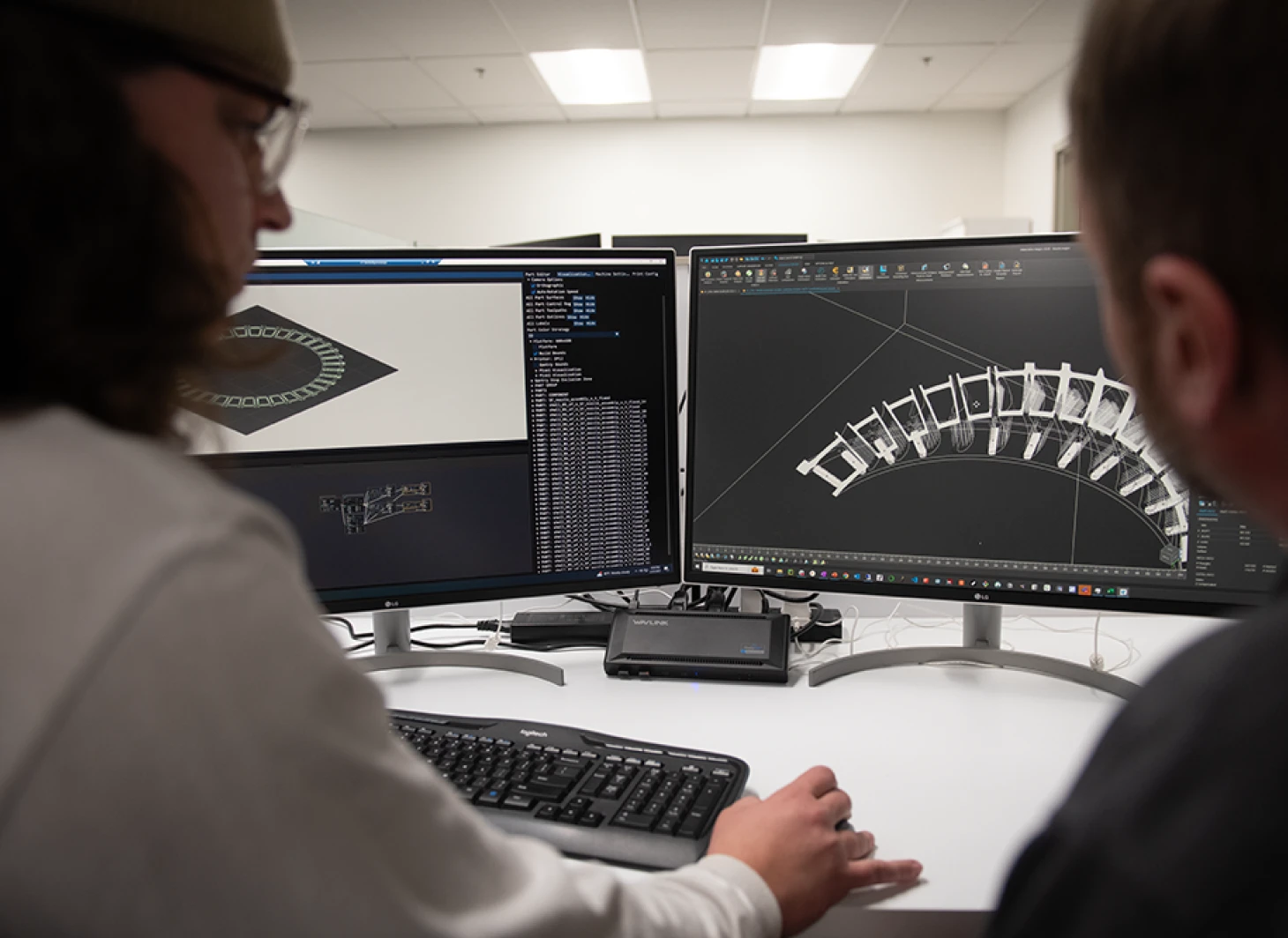 Two professionals collaborate at computers, reviewing 3D design models on multiple screens in a modern workspace.